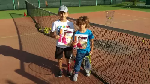 Doi copii fericiți țin cupe și diplome pe terenul de tenis, după o competiție reușită