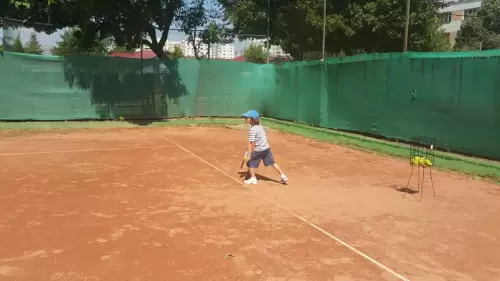 Copil pe terenul de tenis, lângă un coș de mingi, practicând serviciul în timpul antrenamentului