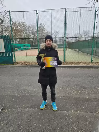 Copil fericit ținând o cupă și o diplomă, cu terenul de tenis în fundal, după competiție