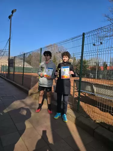Doi copii fericiți țin cupe și diplome pe terenul de tenis, după o competiție reușită